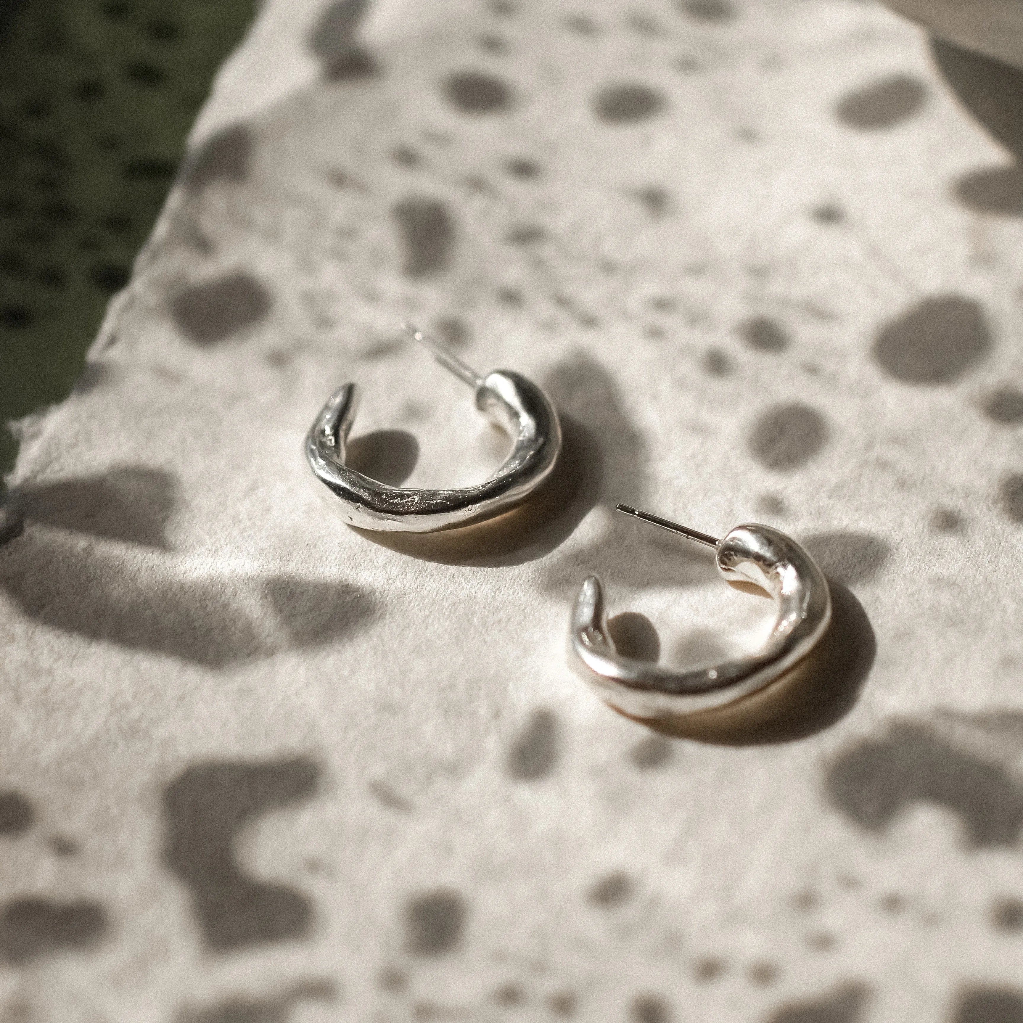 A pair of polished silver hoop earrings resting on a textured white surface with a mottled dark green background and a ceramic bowl in the upper right.