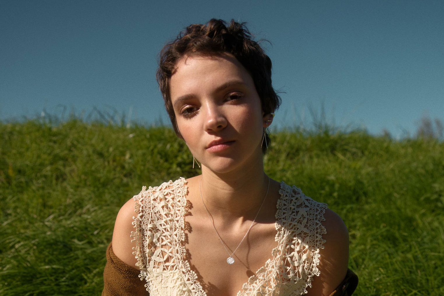 Woman in a white dress and brown cardigan sitting on grass with a clear blue sky.
