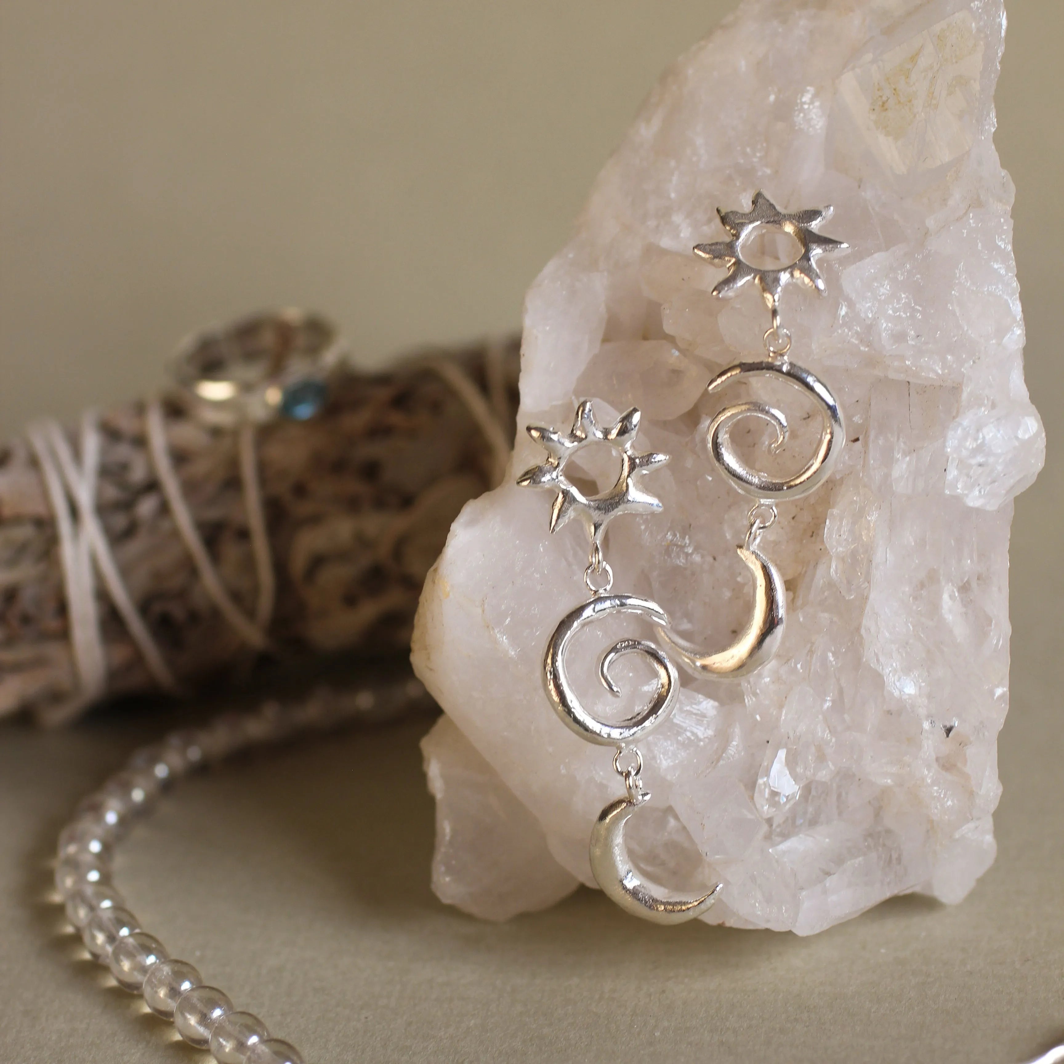sun and moon dangle earrings on a white crystal with bead necklace with blurred sage and ring in the background