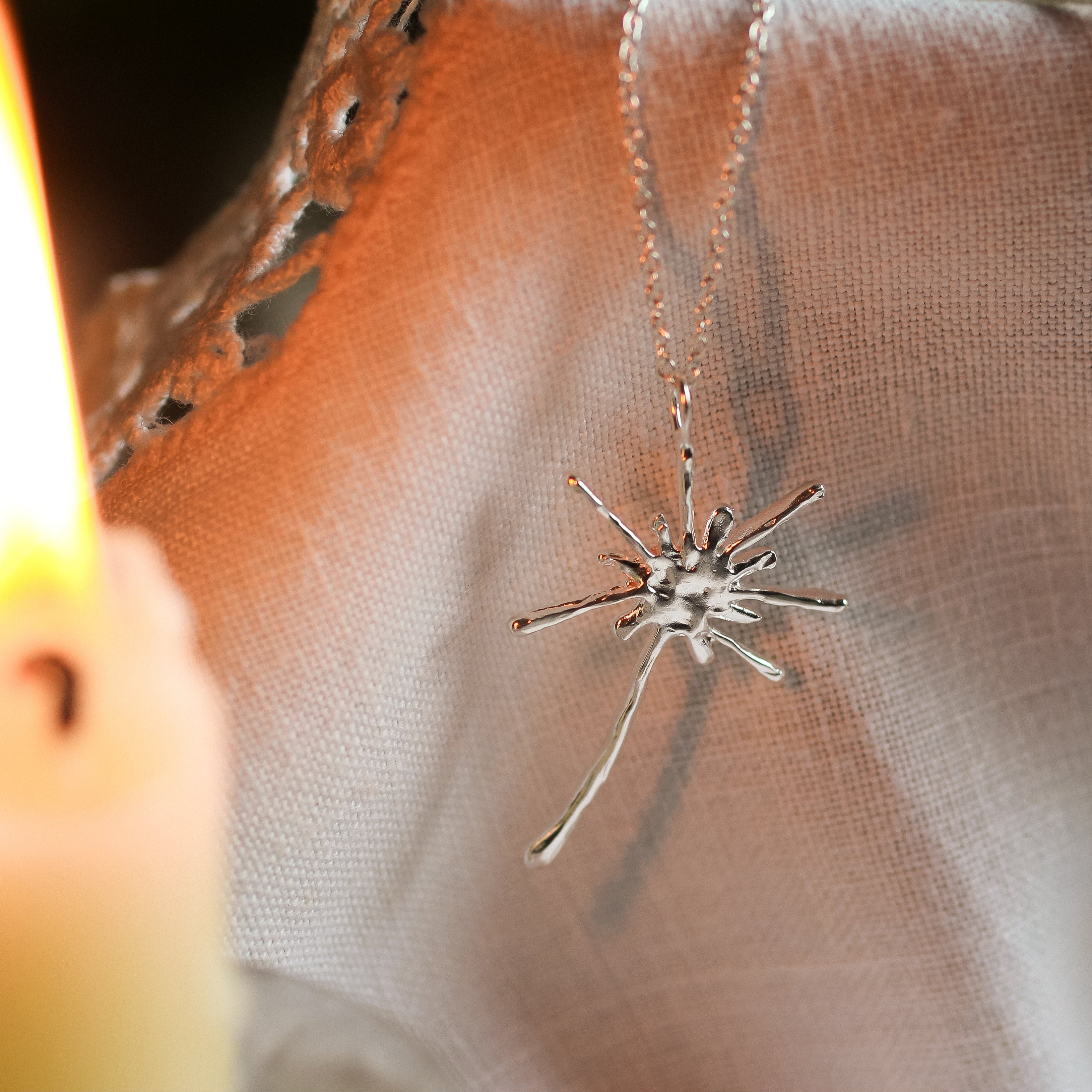 dandelion necklace in silver on neutral fabric with a blurred candle in the foreground