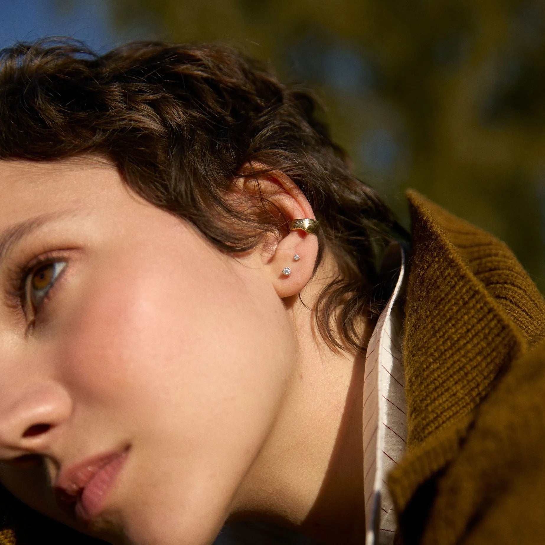 Close-up of a person wearing 2mm and 3mm diamond studs, a brown jacket with a blurred natural background
