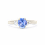 A three-stone diamond ring featuring a central oval-cut blue sapphire with round-cut diamonds on either side, set in a polished white gold band against a plain white background.