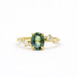 blue green oval sapphire engagement ring with scattered round diamonds on a gold band against a white background