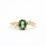 blue green oval sapphire engagement ring with scattered round diamonds on a gold band against a white background