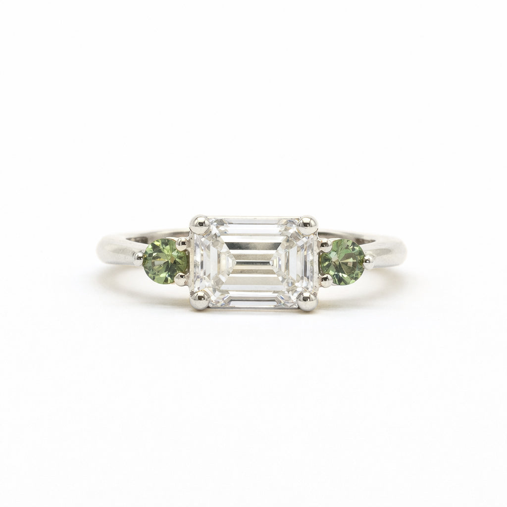A silver-toned white gold three-stone ring featuring a central emerald-cut clear diamond flanked by two round green gemstones, set against a plain white background.