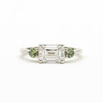 A silver-toned white gold three-stone ring featuring a central emerald-cut clear diamond flanked by two round green gemstones, set against a plain white background.