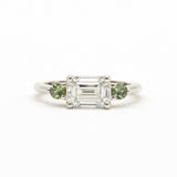 A silver-toned white gold three-stone ring featuring a central emerald-cut clear diamond flanked by two round green gemstones, set against a plain white background.