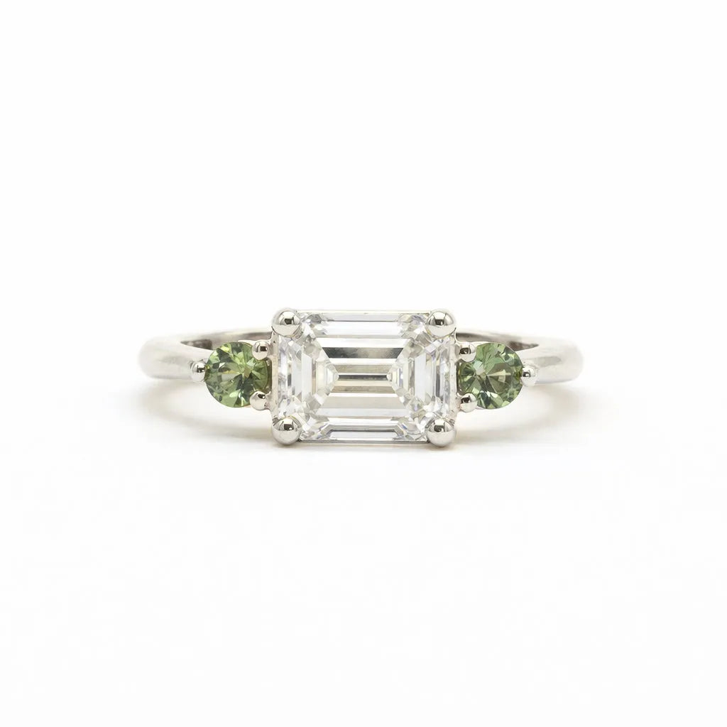 A silver-toned white gold three-stone ring featuring a central emerald-cut clear diamond flanked by two round green gemstones, set against a plain white background.