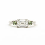A silver-toned white gold three-stone ring featuring a central emerald-cut clear diamond flanked by two round green gemstones, set against a plain white background.