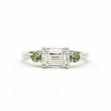 A silver-toned white gold three-stone ring featuring a central emerald-cut clear diamond flanked by two round green gemstones, set against a plain white background.