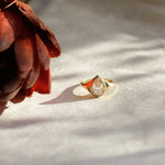 A gold ring with a pear-cut diamond sits on a textured white surface next to a dried reddish-brown flower, lit by dramatic side lighting.