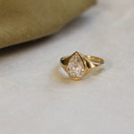 A gold ring featuring a pear-cut clear gemstone, resting on a light beige textured surface with a soft olive-green fabric prop in the background.