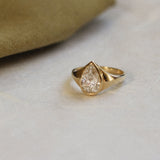 A gold ring featuring a pear-cut clear gemstone, resting on a light beige textured surface with a soft olive-green fabric prop in the background.