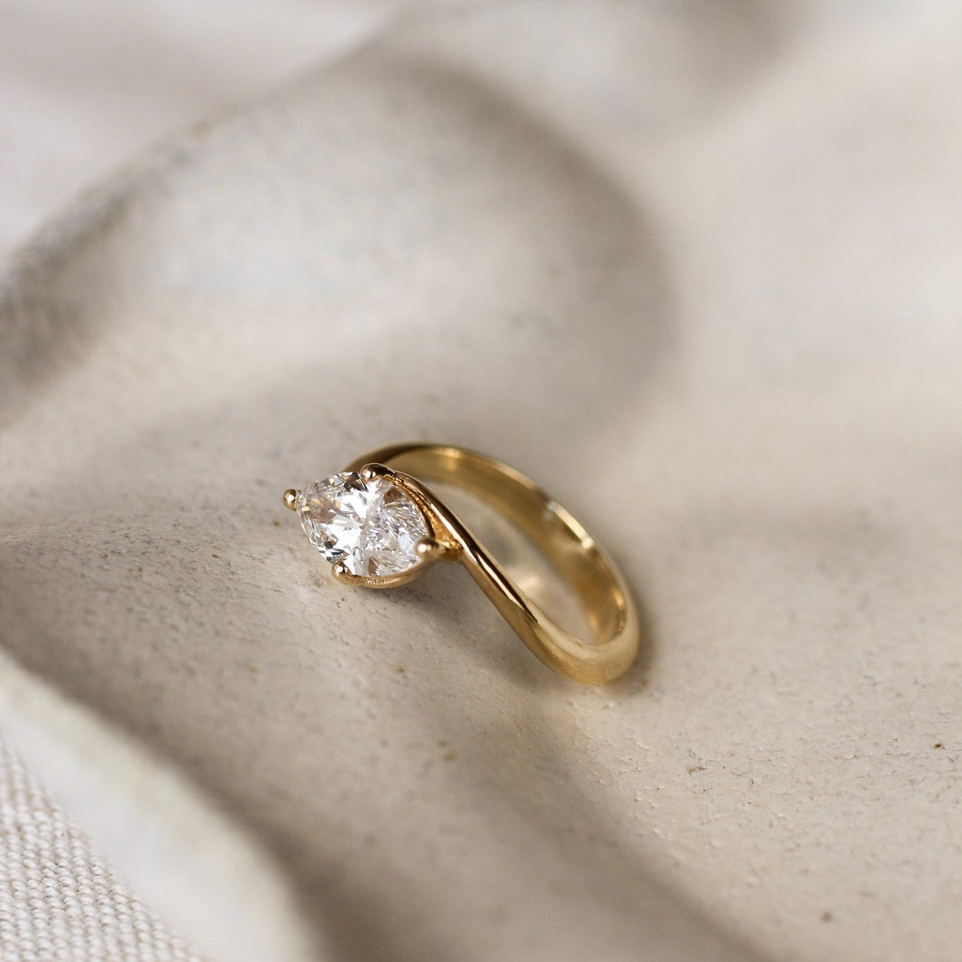 Gold ring with a marquise almond-shaped diamond and curved shank on a beige ceramic dish