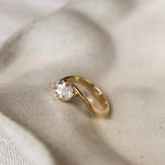 Gold ring with a marquise almond-shaped diamond and curved shank on a beige ceramic dish
