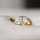 Gold curved engagement ring with a marquise diamond on a white ceramic dish
