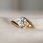 Gold engagement ring with a round diamond and curved wave shank on a textured surface