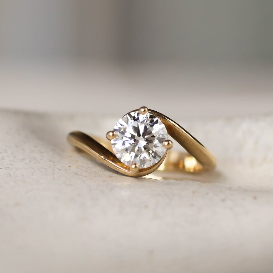 Gold engagement ring with a round diamond and curved wave shank on a textured surface