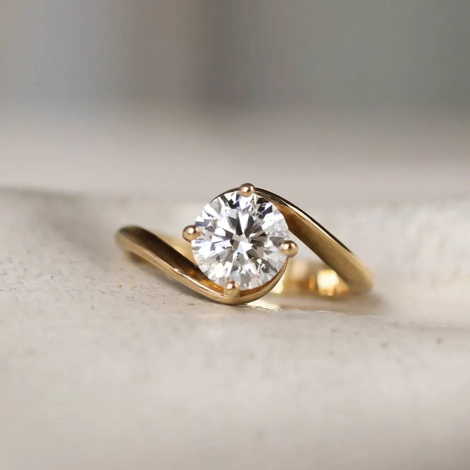 Gold engagement ring with a round diamond and curved wave shank on a textured surface