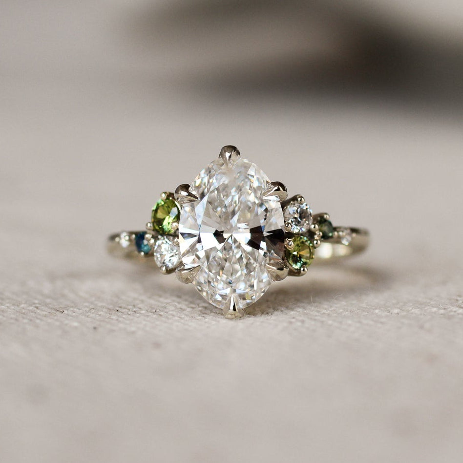 Large oval diamond ring with small green and blue gemstones on a textured surface
