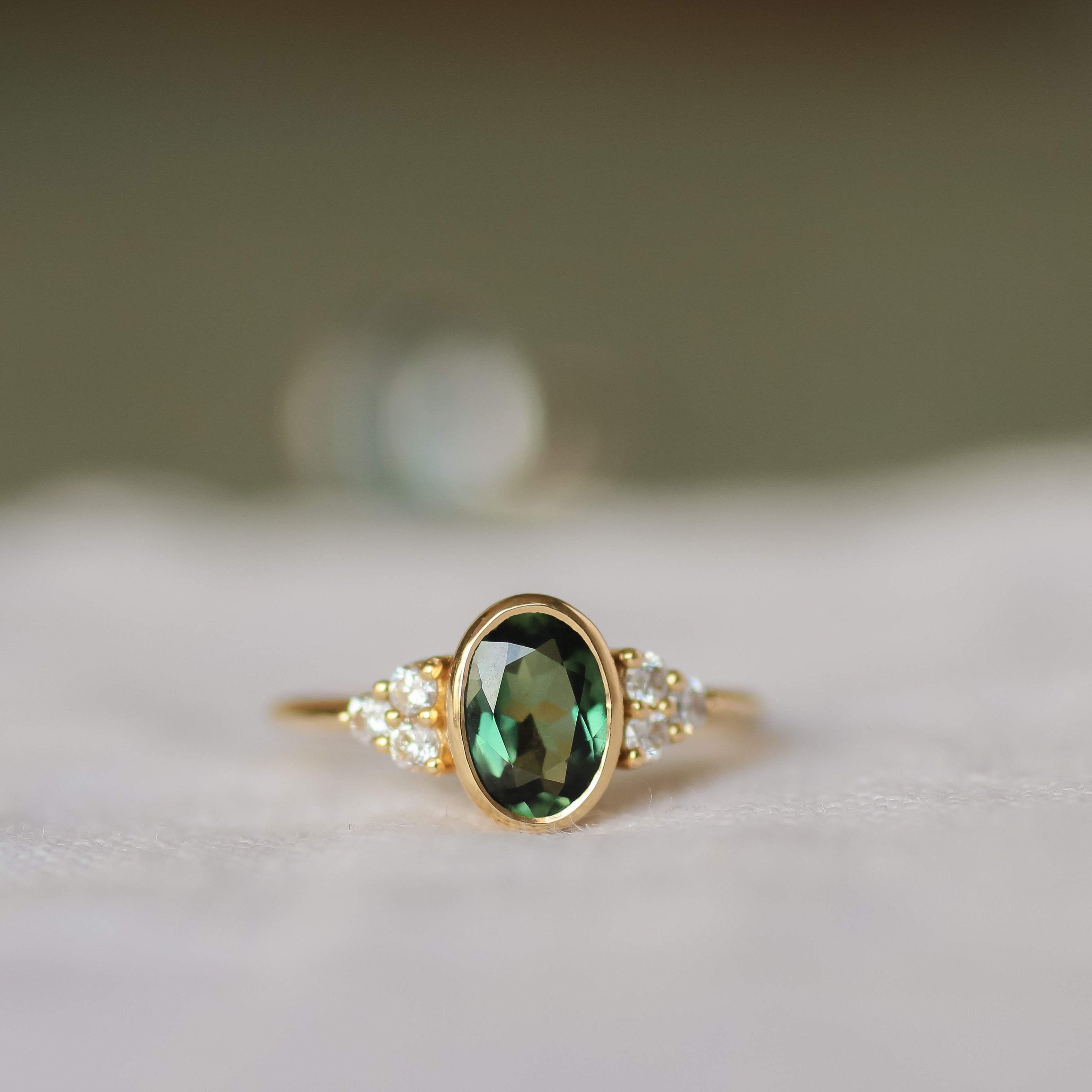 gold ring with oval bezel set sapphire and a cluster of 3 round diamonds either side with a blurred background