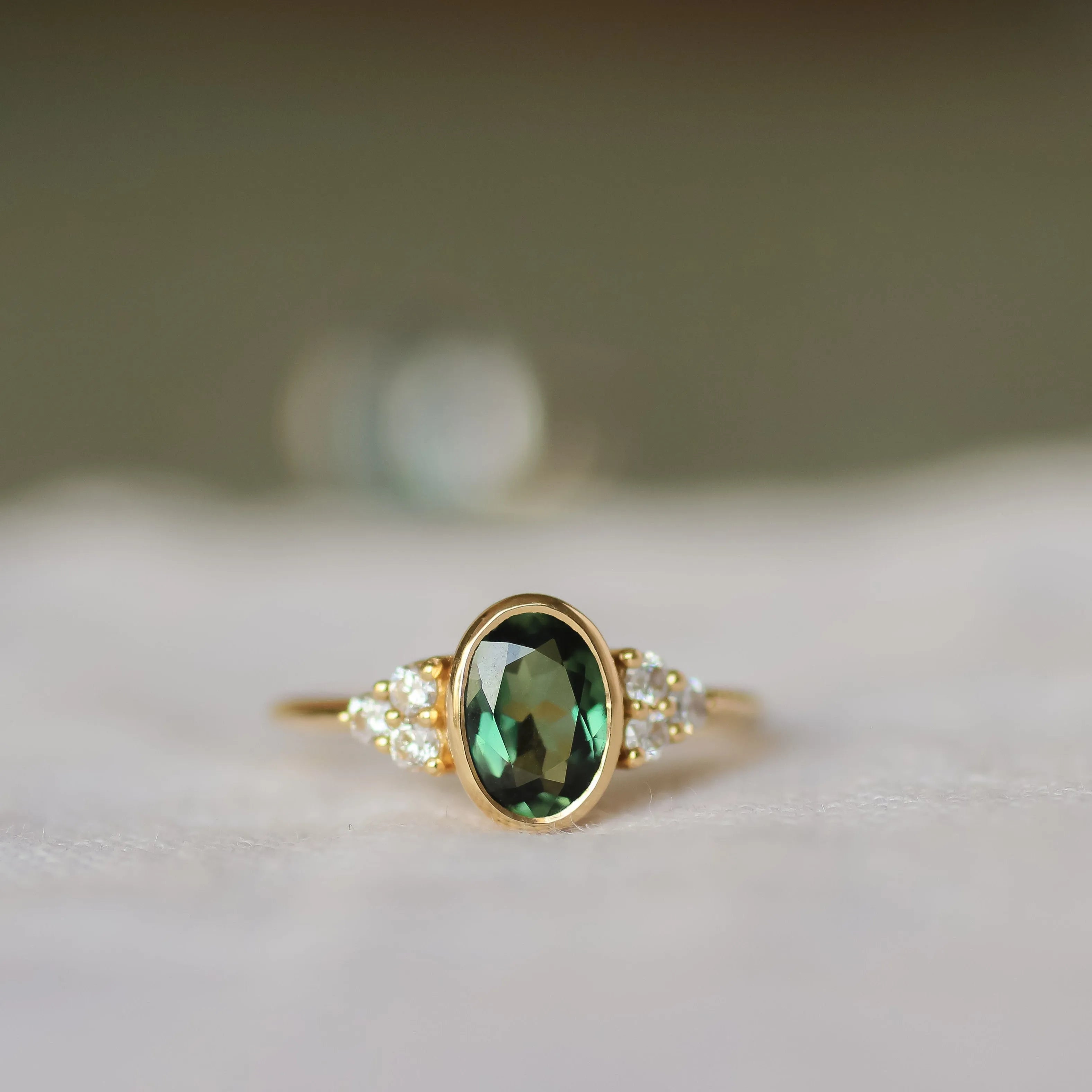 gold ring with oval bezel set sapphire and a cluster of 3 round diamonds either side with a blurred background