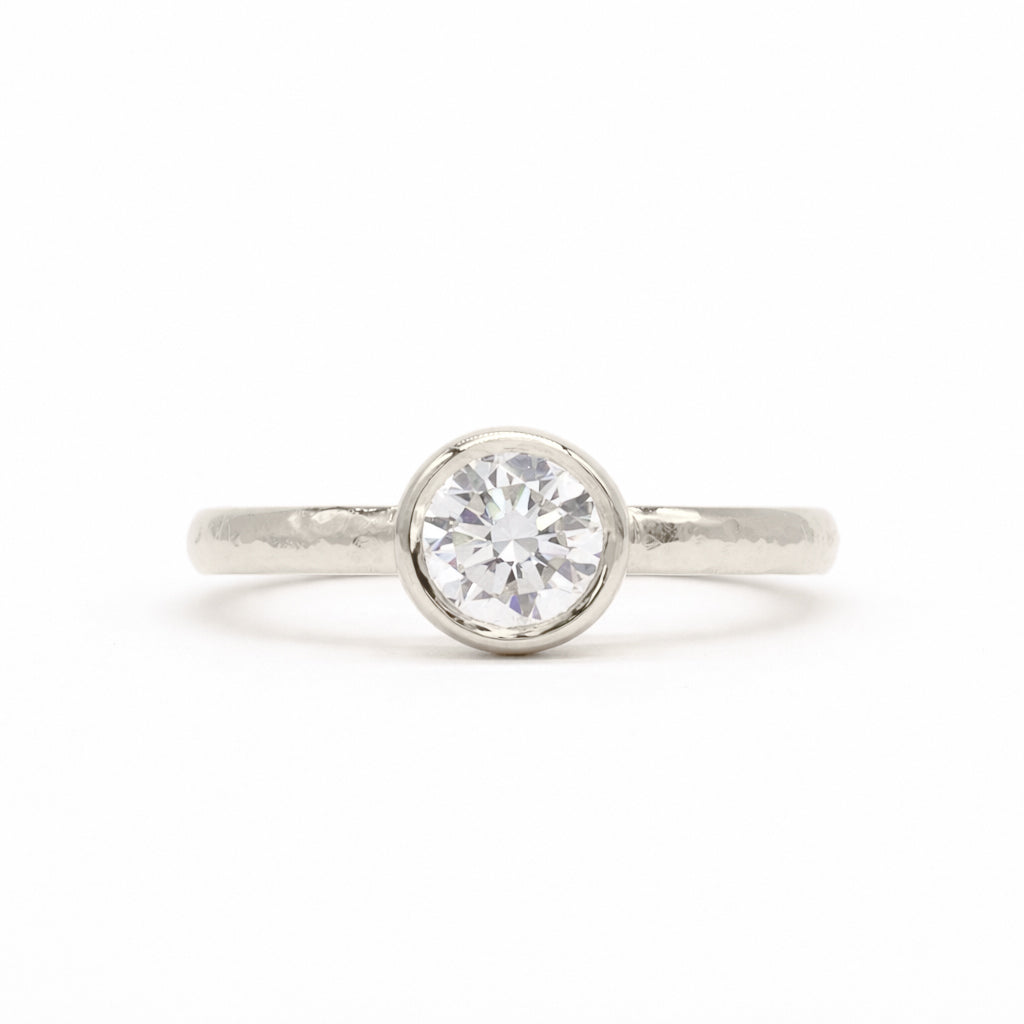 A white-gold solitaire engagement ring with a round brilliant-cut diamond set in a bezel setting, shown on a clean white background.