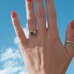 Hand with a blue sapphire and diamond bezel engagement ring against a blue sky