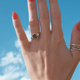 Hand with a blue sapphire and diamond bezel engagement ring against a blue sky
