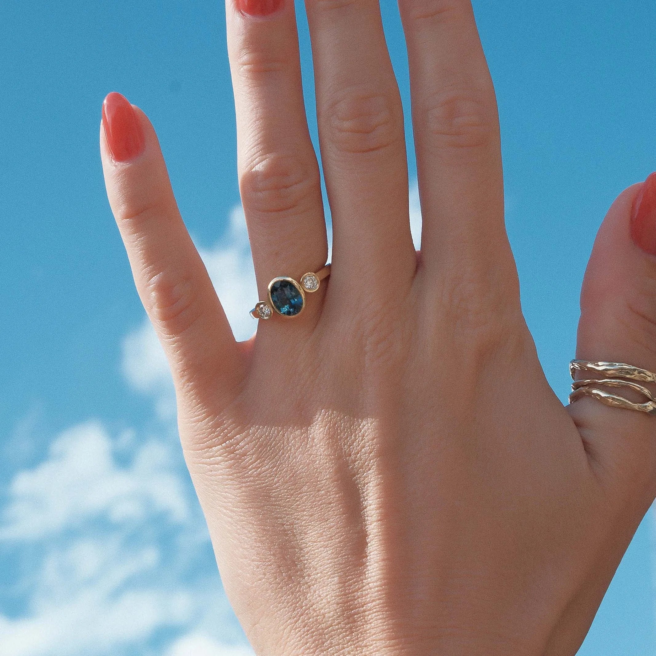Hand with a blue sapphire and diamond bezel engagement ring against a blue sky