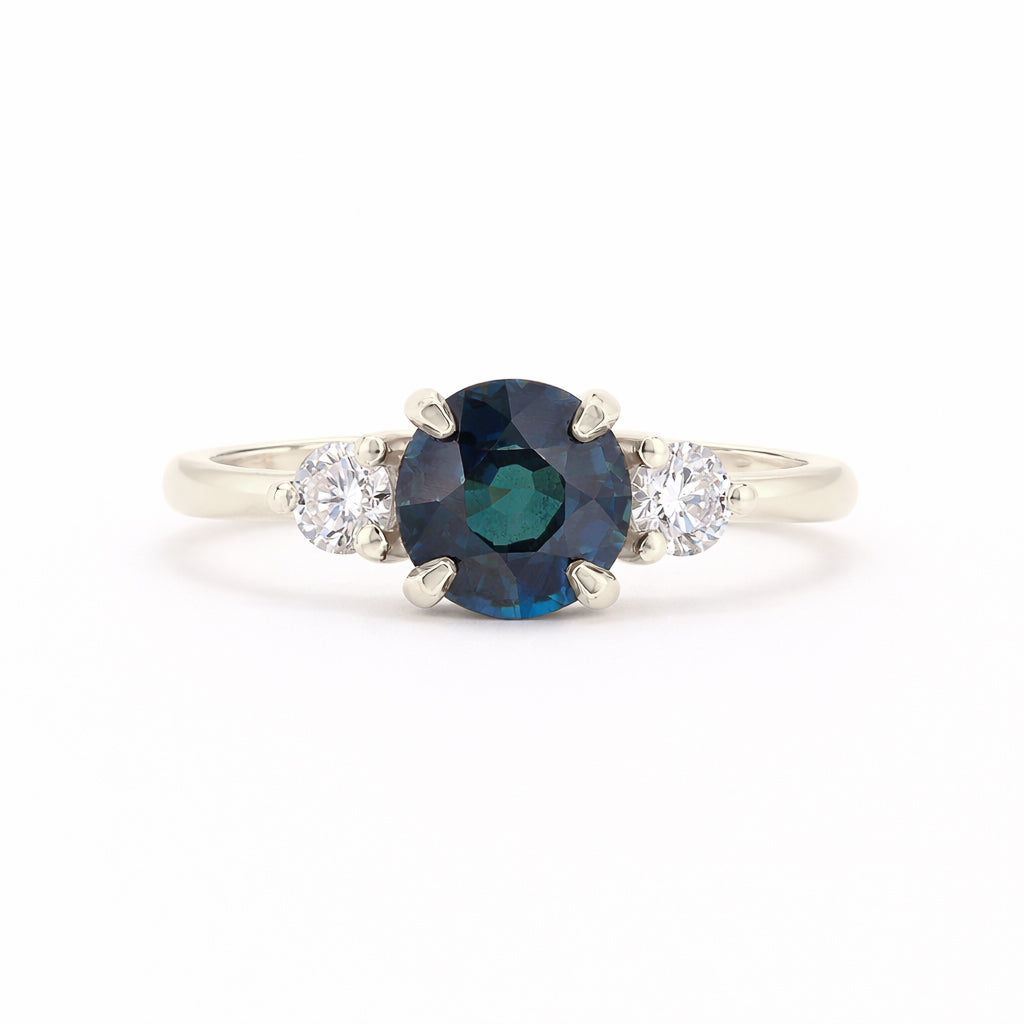 A silver-tone ring featuring a round-cut dark blue sapphire center stone flanked by two small round-cut clear diamonds on a white background.