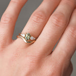 Close-up of a hand wearing a gold engagement with a green gemstone and diamonds with a curved wedding ring on a neutral background