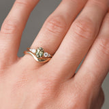 Close-up of a hand wearing a gold engagement with a green gemstone and diamonds with a curved wedding ring on a neutral background