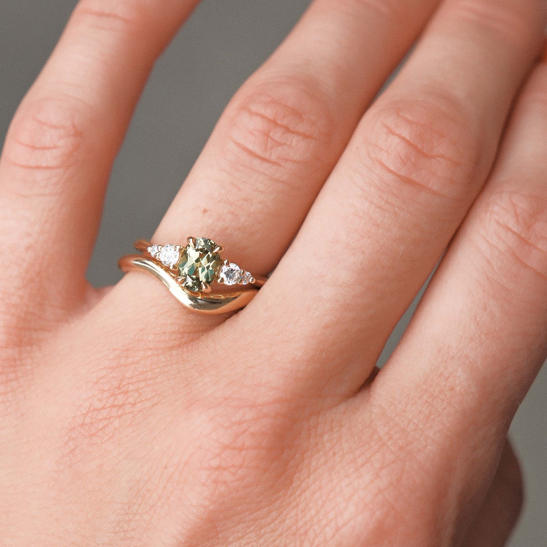 Close-up of a hand wearing a gold engagement with a green gemstone and diamonds with a curved wedding ring on a neutral background