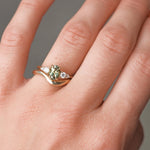 Close-up of a hand wearing a gold engagement with a green gemstone and diamonds with a curved wedding ring on a neutral background