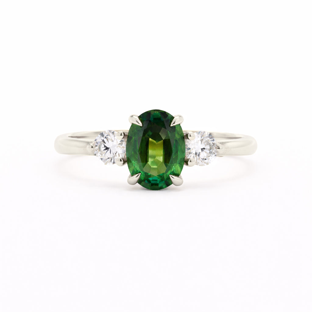 A silver-toned white-gold three-stone ring featuring a central oval-cut green gemstone flanked by two round clear stones, set against a plain white background.