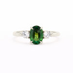 A silver-toned white-gold three-stone ring featuring a central oval-cut green gemstone flanked by two round clear stones, set against a plain white background.
