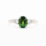 A silver-toned white-gold three-stone ring featuring a central oval-cut green gemstone flanked by two round clear stones, set against a plain white background.