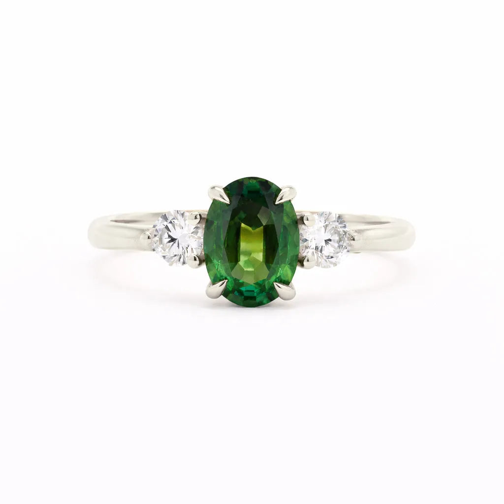 A silver-toned white-gold three-stone ring featuring a central oval-cut green gemstone flanked by two round clear stones, set against a plain white background.