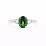 A silver-toned white-gold three-stone ring featuring a central oval-cut green gemstone flanked by two round clear stones, set against a plain white background.