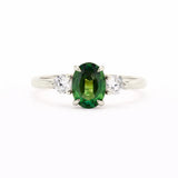 A silver-toned white-gold three-stone ring featuring a central oval-cut green gemstone flanked by two round clear stones, set against a plain white background.