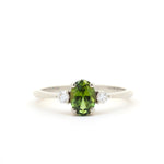 A white-gold three-stone ring featuring a central oval-cut green peridot gemstone flanked by two small round-cut clear diamonds, set against a plain white background.