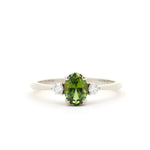 A white-gold three-stone ring featuring a central oval-cut green peridot gemstone flanked by two small round-cut clear diamonds, set against a plain white background.