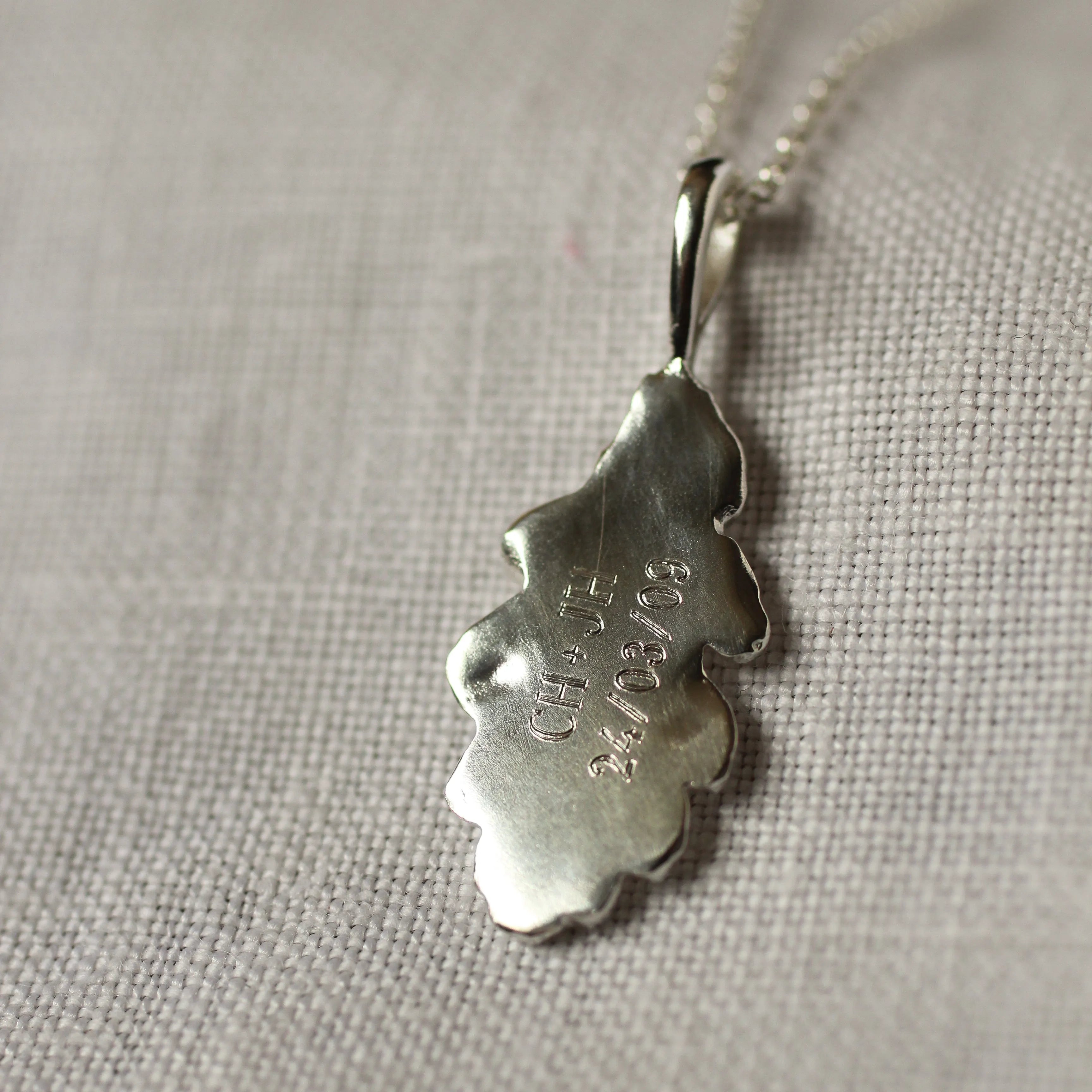 Silver leaf-shaped pendant with 'CH + JH 24/03/09' engraved on a chain against a neutral background