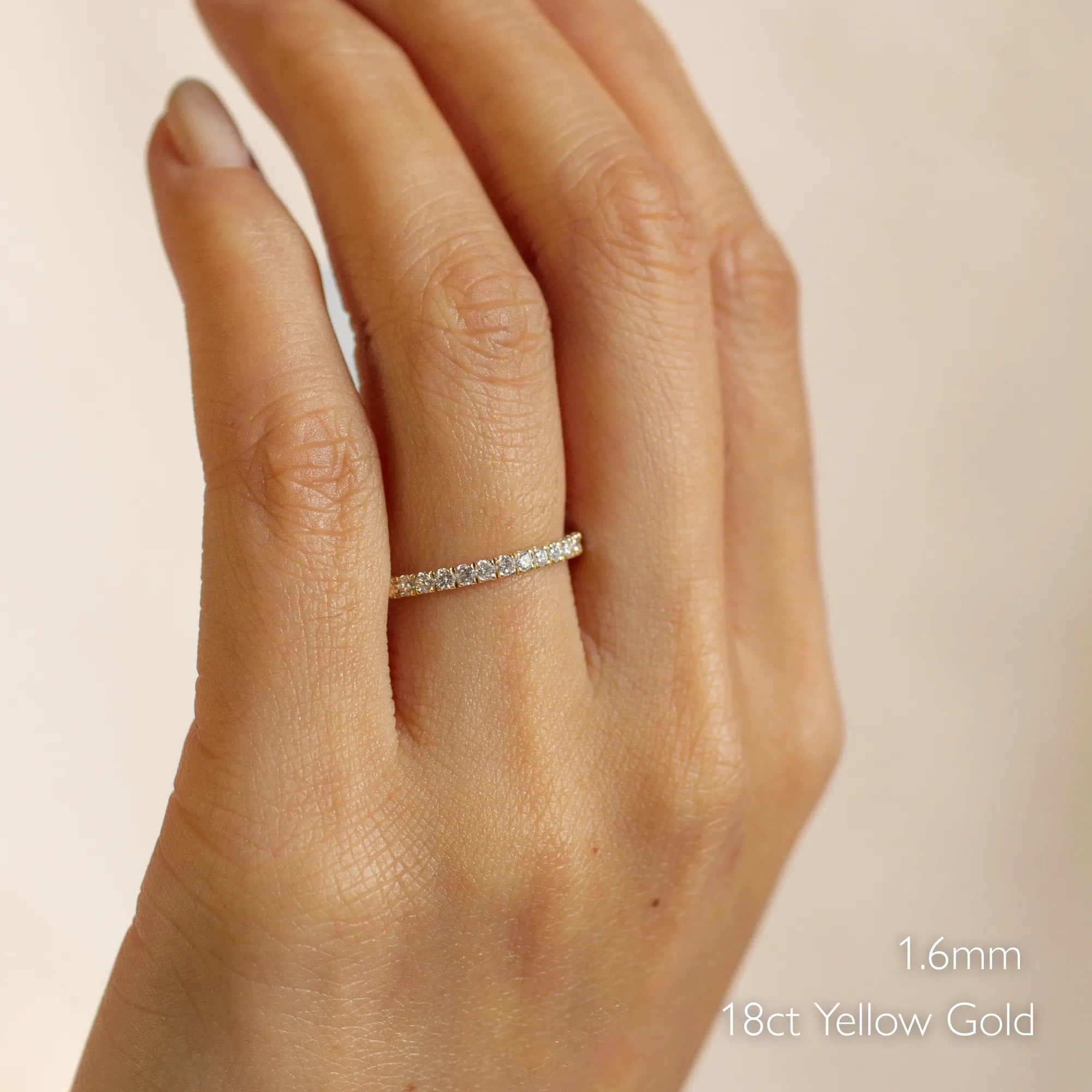 Close-up of a hand wearing a 1.6mm diamond wedding ring with '18ct Yellow Gold' text on a beige background