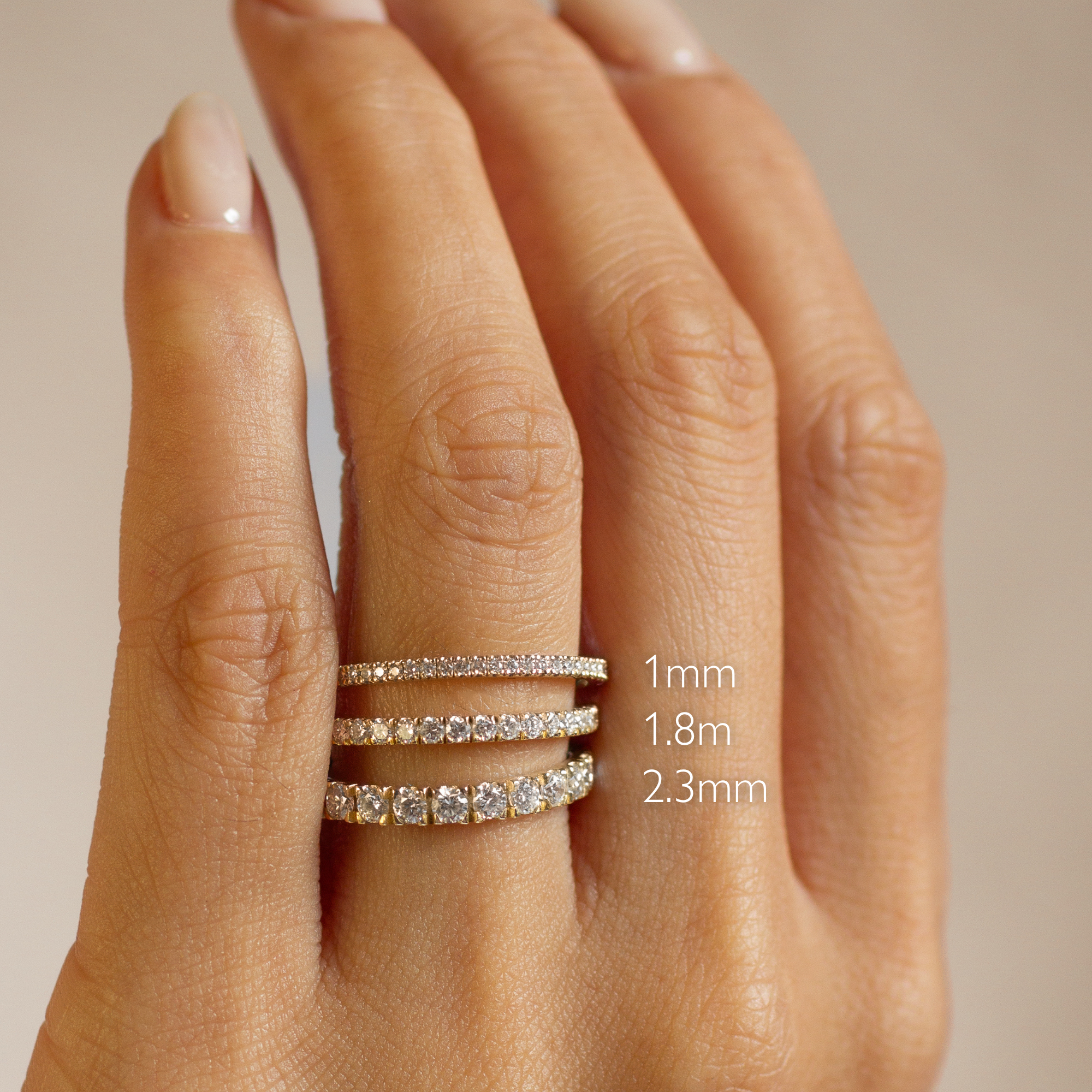 A close-up of a hand wearing three slim diamond bands on the ring finger, with text indicating widths: 1mm, 1.8mm, and 2.3mm.