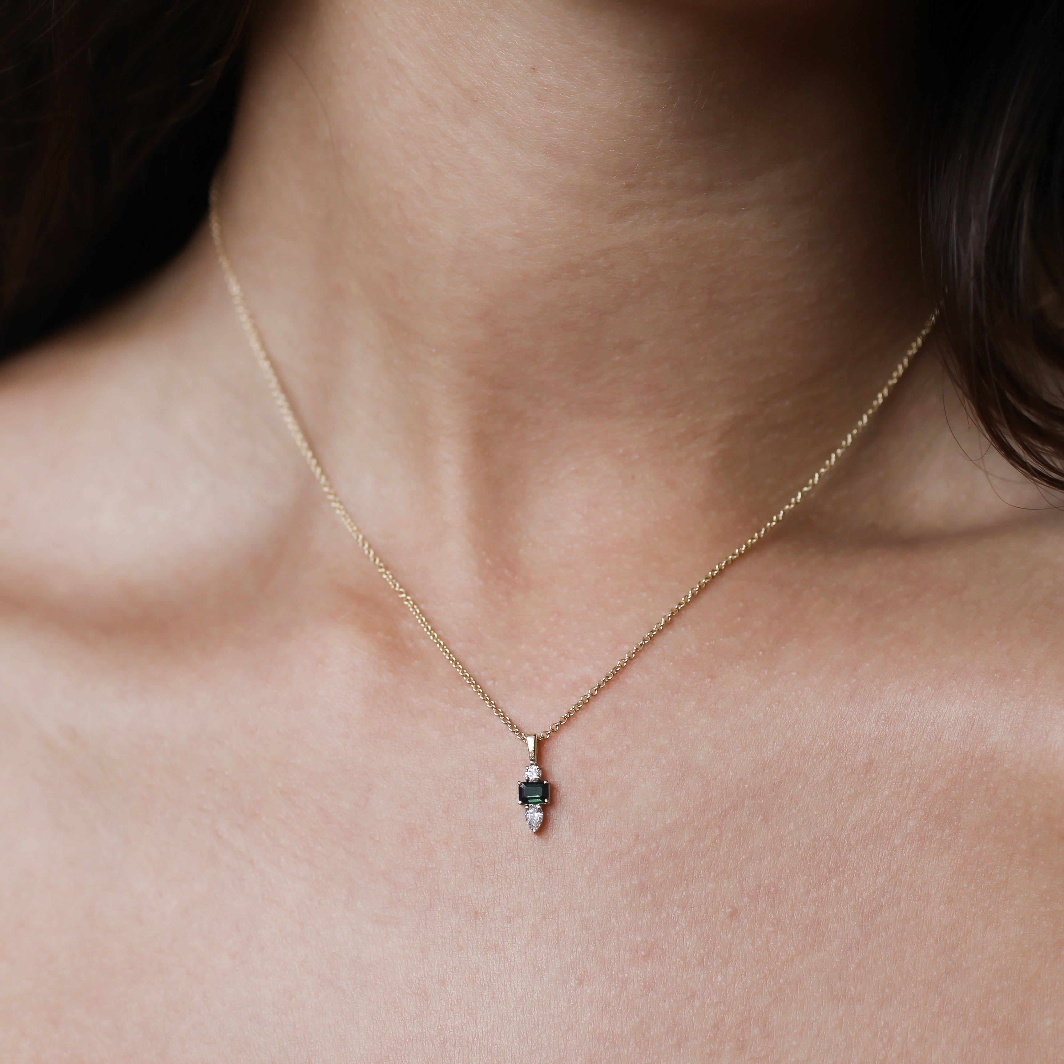 A close-up of a delicate gold chain necklace with a small teardrop-shaped pendant featuring a dark green stone, worn on a model's neck.