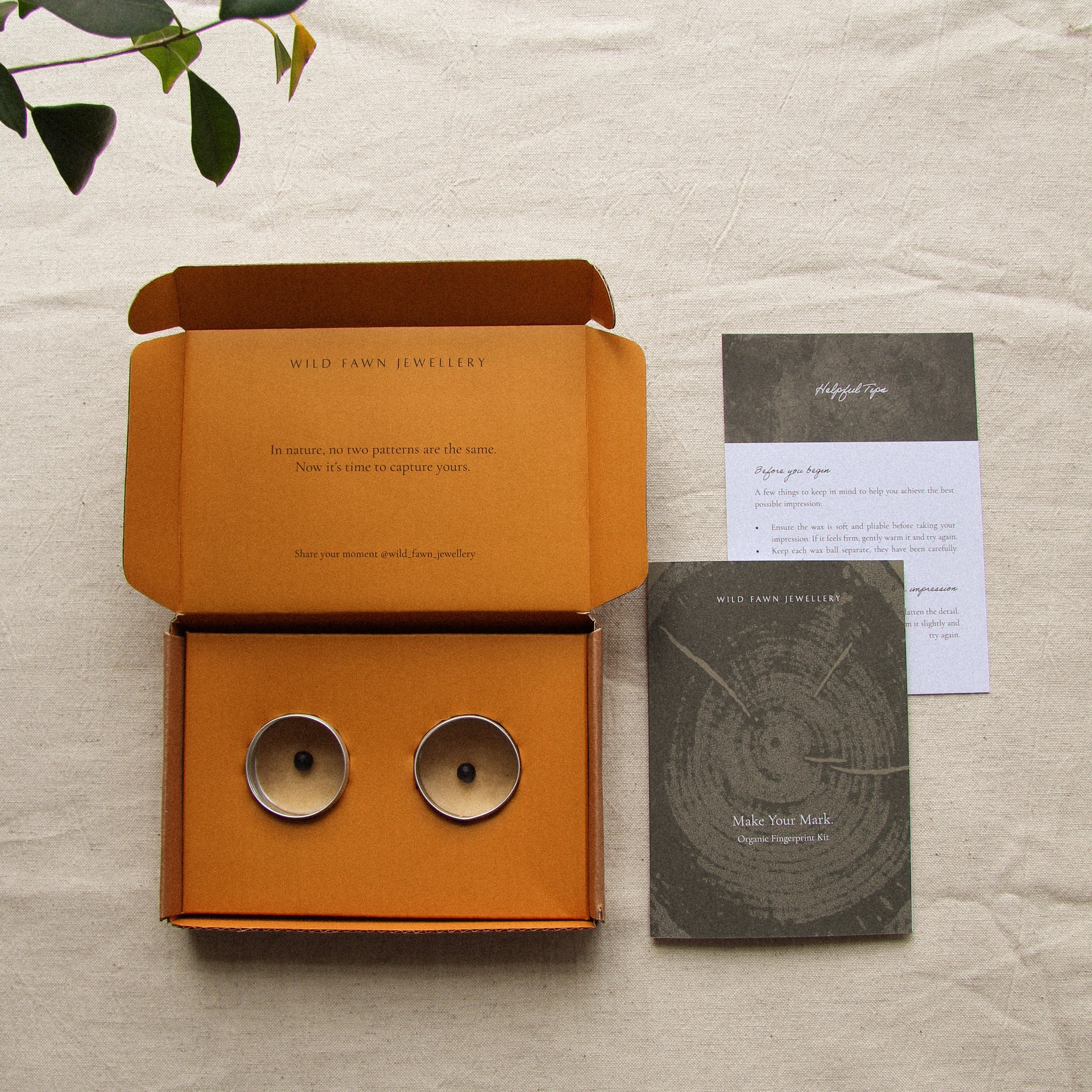 Orange box with two metallic tins with balls of imprint wax and an instructional card on a beige surface with a plant.