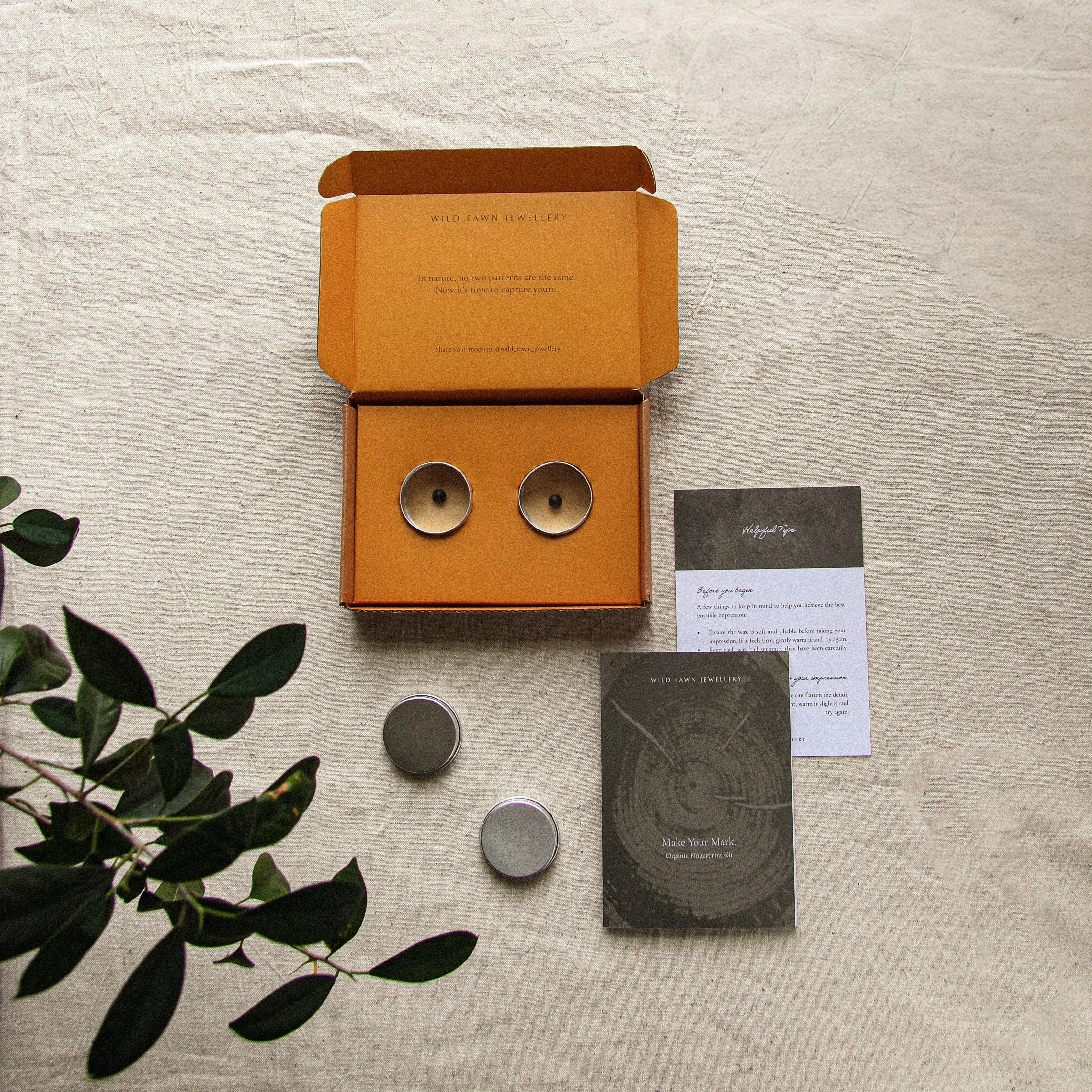 Orange box with two round tins with jewellery imprint wax and an instructional card on a textured surface with green leaves.