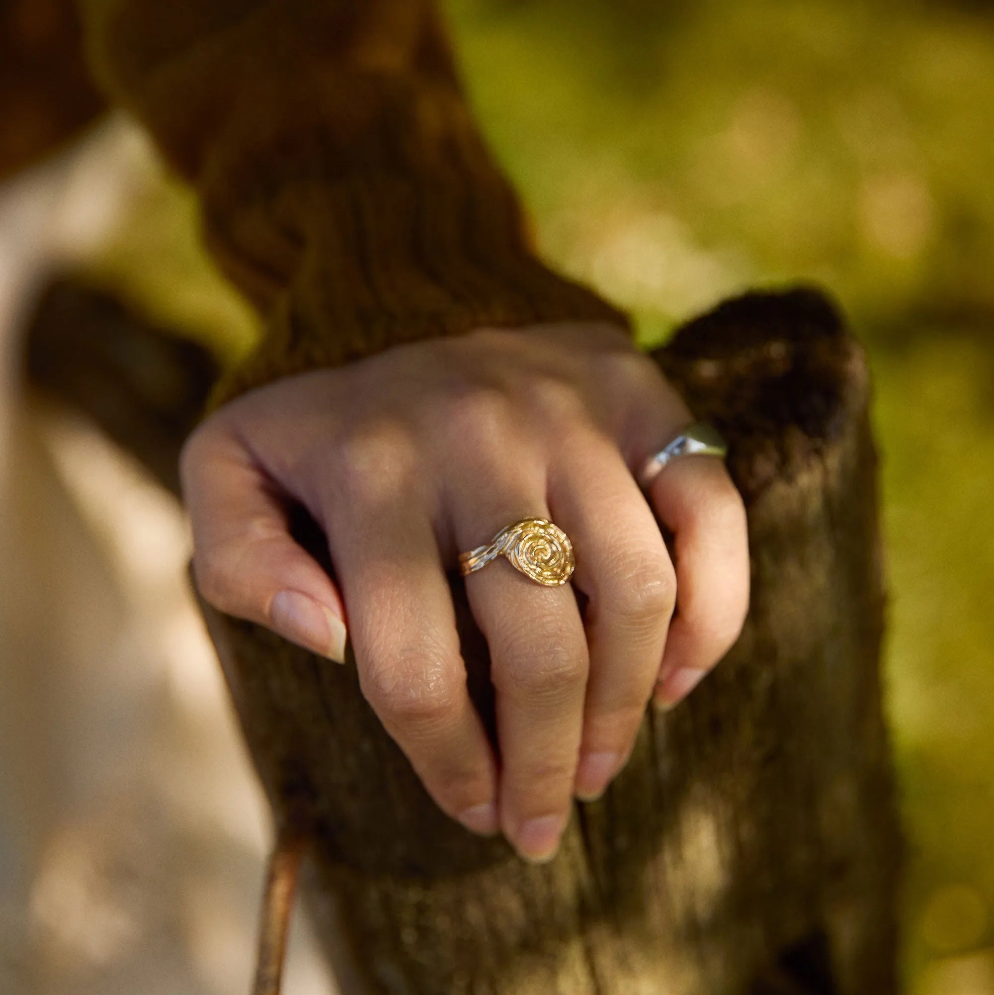 Hand leaning on wooden fence wearing a gold swirl ring on a blurred natural background